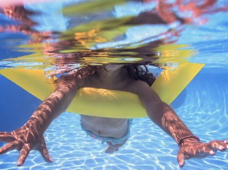 Underwater view of a girl swimming towards the camera with her yellow float in the pool, summer sunlight reflected on the bottom of the pool and there are also reflections on the surface of the water