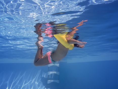 underwater view of a child swimming relaxed with his yellow float in the pool, summer sunlight reflected on the blue water surface