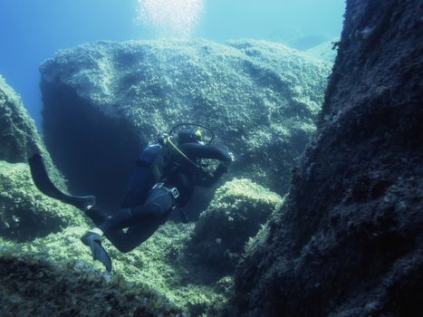 Diver passes close to the seabed of rocks covered in small algae, the bubbles of his breath rise through the crystalline blue and turquoise water illuminated by the sun