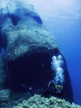 A person dives near the seabed next to a huge rock known as Pietra di Saragossa. The bubbles of the diver's breathing rise to the surface illuminated by sunlight. Small fish swim next to the stone