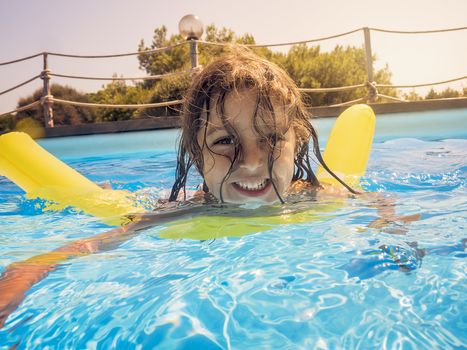 child smiles cheerfully as she swims in the blue water of the pool with her yellow float, behind her the glow of the summery sun gleams golden