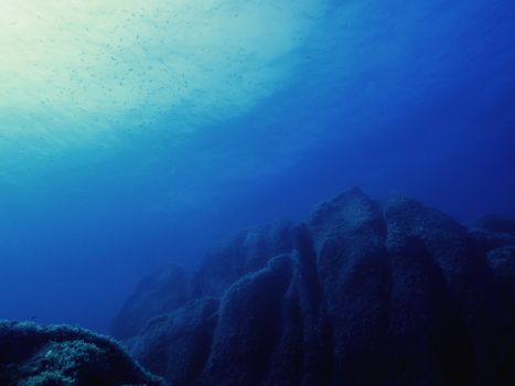 large rocks of the seabed lit up slightly by the sun reflected on the surface of the sea, small fish swim in crystal clear blue and turquoise water