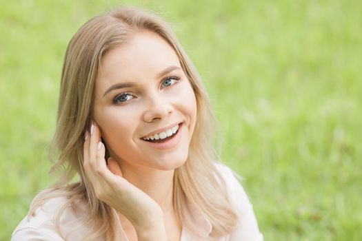 Smiling young blonde woman touching face skin over green natural grass background
