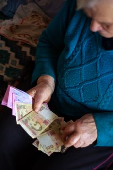 Old poor gray hair woman holds Ukrainian hryvna money in her hands. Woman is sad. Poor life in village. Old age not good. Low-light photo.