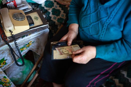 Old poor gray hair woman holds Ukrainian hryvna money in her hands. Woman is sad. Poor life in village. Old age not good. Low-light photo.