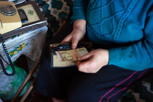 Old poor gray hair woman holds Ukrainian hryvna money in her hands. Woman is sad. Poor life in village. Old age not good. Low-light photo.