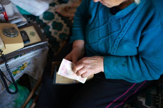 Old poor gray hair woman holds Ukrainian hryvna money in her hands. Woman is sad. Poor life in village. Old age not good. Low-light photo.