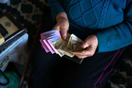 Old poor gray hair woman holds Ukrainian hryvna money in her hands. Woman is sad. Poor life in village. Old age not good. Low-light photo.