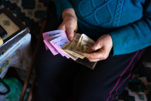 Old poor gray hair woman holds Ukrainian hryvna money in her hands. Woman is sad. Poor life in village. Old age not good. Low-light photo.