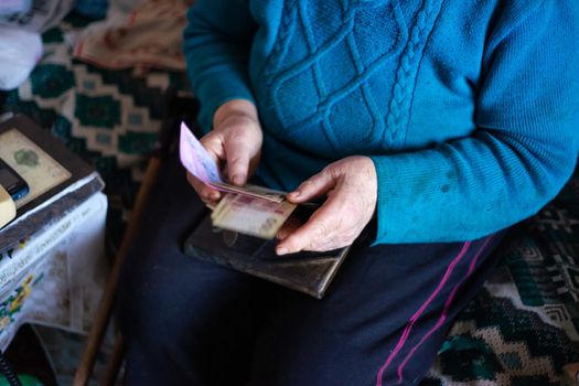 Old poor gray hair woman holds Ukrainian paper money in her hands. Woman is sad. Poor life in village. Old age not good. Low-light photo.