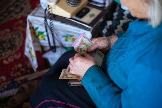 Old poor gray hair woman holds Ukrainian paper money in her hands. Woman is sad. Poor life in village. Old age not good. Low-light photo.