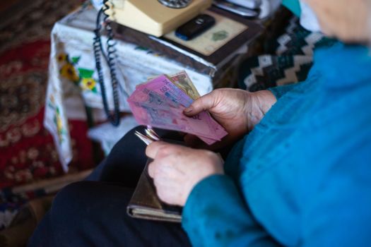 Old poor gray hair woman holds Ukrainian paper money in her hands. Woman is sad. Poor life in village. Old age not good. Low-light photo.