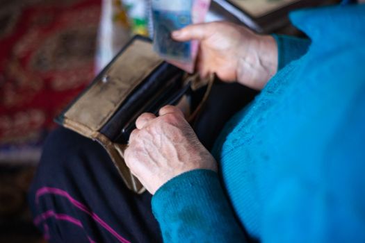 Old poor gray hair woman putting paper Ukrainian money in her vintage leather wallet by her hands. Woman is sad. Poor life in village. Old age not good. Low-light photo.