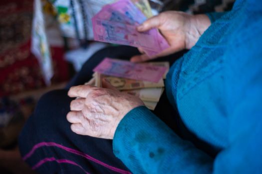 Old poor gray hair woman holds Ukrainian paper money in her hands. Woman is sad. Poor life in village. Old age not good. Low-light photo.