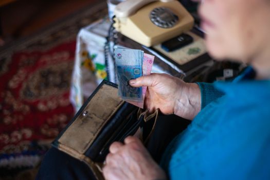 Old poor gray hair woman putting paper Ukrainian money in her vintage leather wallet by her hands. Woman is sad. Poor life in village. Old age not good. Low-light photo.