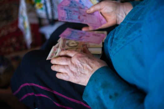Old poor gray hair woman holds Ukrainian paper money in her hands. Woman is sad. Poor life in village. Old age not good. Low-light photo.