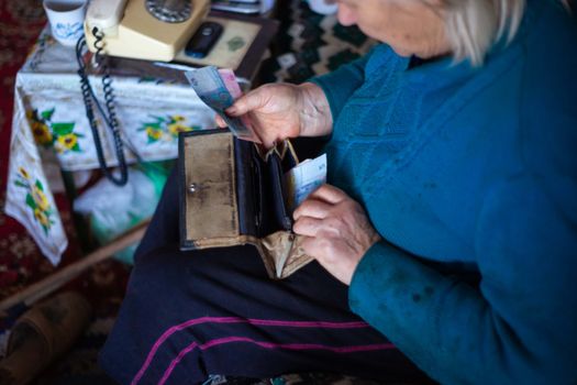 Old poor gray hair woman putting paper Ukrainian money in her vintage leather wallet by her hands. Woman is sad. Poor life in village. Old age not good. Low-light photo.