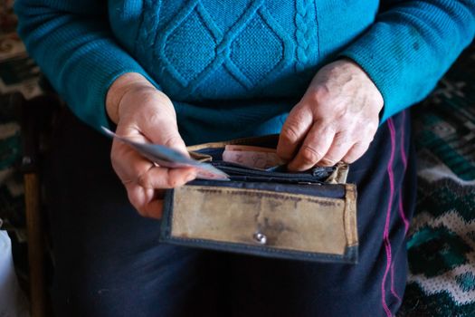 Old poor gray hair woman putting paper Ukrainian money in her vintage leather wallet by her hands. Woman is sad. Poor life in village. Old age not good. Low-light photo.