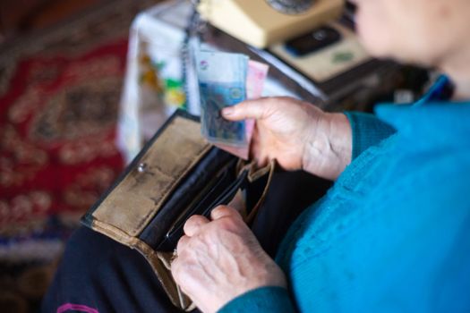 Old poor gray hair woman putting paper Ukrainian money in her vintage leather wallet by her hands. Woman is sad. Poor life in village. Old age not good. Low-light photo.
