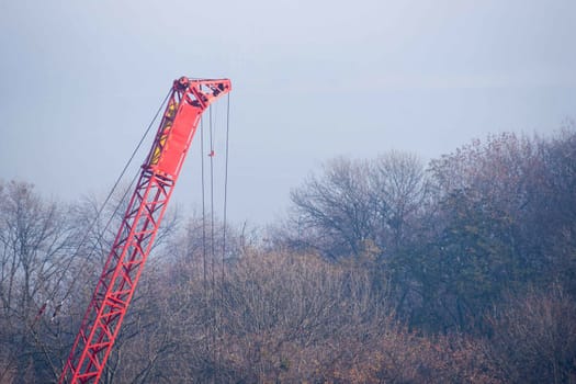 Construction process with construction automotive crane with red gibbet. Autumn fog and river bridge  on blurred background. Forest around crane.