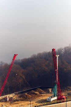 Construction process with construction automotive cranes with red and white gibbets. Autumn fog and river bridge  on blurred background. Forest around cranes.