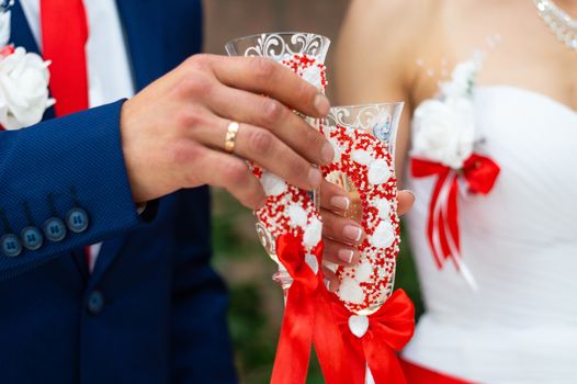 Bride in white dress and groom in dark blue suit clink glasses with beautiful handmade glasses at the wedding table.