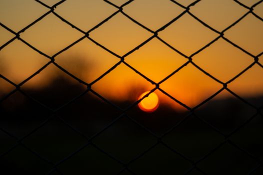 Orange evening sky with blurred sun on horizon trough fence. Creative idea- underexposed photo.