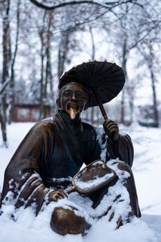 Bronze monument of a chinese old man with umbrella in winter snow park. Closeup with blurred background.