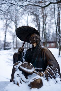 Bronze monument of a chinese old man with umbrella in winter snow park. Closeup with blurred background.
