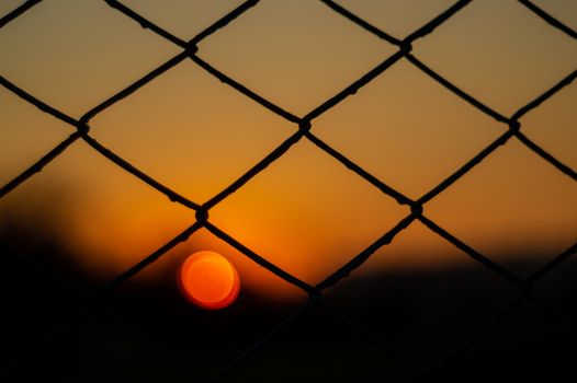 Orange evening sky with blurred sun on horizon trough fence. Creative idea- underexposed photo.