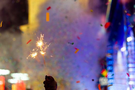 New Year celebration on main square. A lot of smoke or fog, lights, salute and confetti. Blurred colorful background. Bengali fire in hand with glove on foreground. Concert scene with blue light at right side of frame.