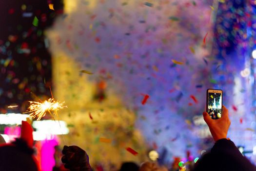 New Year celebration on main square. A lot of smoke or fog, lights, salute and confetti. Blurred colorful background. Bengali fire in hand with glove on foreground. Phone with recorder in other hand. Concert scene with blue light at right side of frame.
