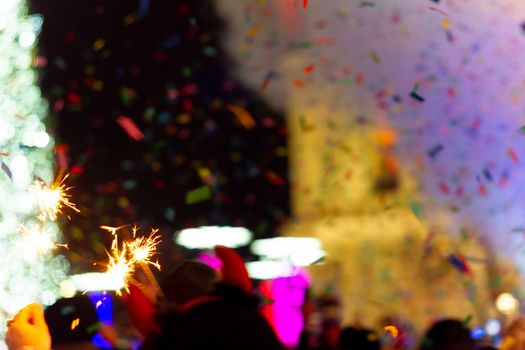 New Year celebration on main square. A lot of smoke or fog, lights, salute and confetti. Blurred colorful background. Bengali fire in hand with glove on foreground. Concert scene with blue light at right side of frame.