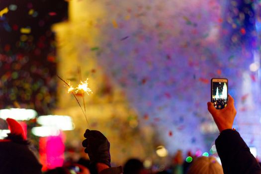 New Year celebration on main square. A lot of smoke or fog, lights, salute and confetti. Blurred colorful background. Bengali fire in hand with glove on foreground. Phone with recorder in other hand. Concert scene with blue light at right side of frame.