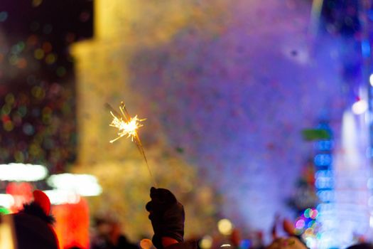 New Year celebration on main square. A lot of smoke or fog, lights, salute and confetti. Blurred colorful background. Bengali fire in hand with glove on foreground. Concert scene with blue light at right side of frame.