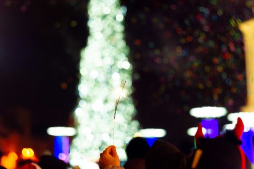 New Year celebration on main square. A lot of smoke or fog, lights, salute and confetti. Blurred colorful background. Bengali fire in hand with glove on foreground. Concert scene with blue light at right side of frame.