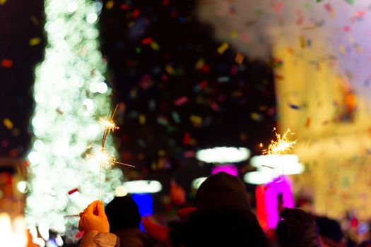 New Year celebration on main square. A lot of smoke or fog, lights, salute and confetti. Blurred colorful background. Bengali fire in hand with glove on foreground. Concert scene with blue light at right side of frame.