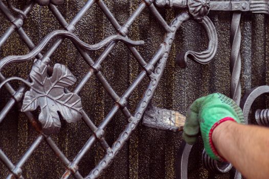 A man paints a gate with forged elements. On his hand is a green glove, near the elbow of a graze. The hand is located on the right of the frame.