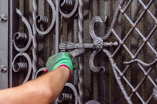 A man paints a gate with forged elements. On his hand is a green glove, near the elbow of a graze. The hand is located on the left of the frame.