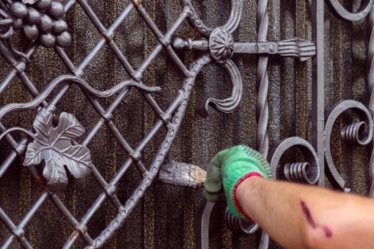 A man paints a gate with forged elements. On his hand is a green glove, near the elbow of a graze. The hand is located on the right of the frame.