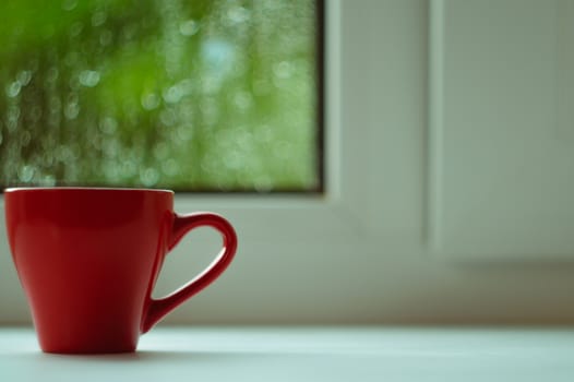 The red espresso cup on white windowsill. On background window and rain. Cup located at left of frame.