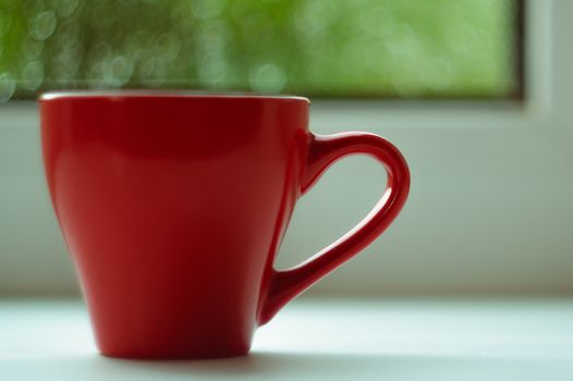 The red espresso cup on white windowsill. On background window and rain. Cup located at left of frame.