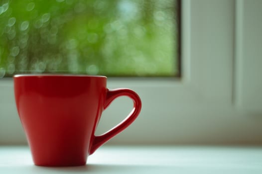 The red espresso cup on white windowsill. On background window and rain. Cup located at left of frame.
