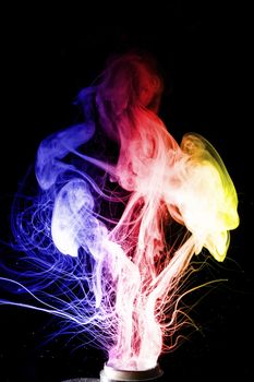 Male hand holds vape. Vape clouds splash from coil on black background. Fog is blue red and yellow colours. Stock isolated smoke with spray boiling glycerine.