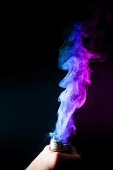 Male hand holds vape. Vape clouds splash from vape coil on black background. Fog is dual color — blue and purple. Stock isolated colorful smoke with spray boiling glycerine.