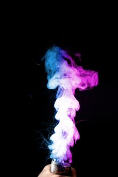 Male hand holds vape. Vape clouds splash from vape coil on black background. Fog is dual color — blue and purple. Stock isolated colorful smoke with spray boiling glycerine.