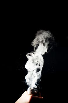 Man hand holds vape. Vape clouds splash from coil on black background. Fog is white. Stock isolated white smoke with spray boiling glycerine around.