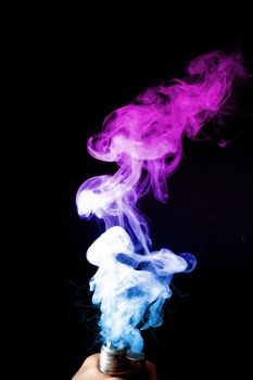 Male hand holds vape. Vape clouds splash from vape coil on black background. Fog is dual color — blue and purple. Stock isolated colorful smoke with spray boiling glycerine.