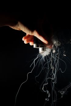 Male hand holds vape upside down. Vape clouds splash from coil on black background. Fog is white. Stock isolated white smoke with spray boiling glycerine.