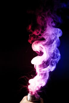 Male hand holds vape. Vape clouds splash from vape coil on black background. Fog is dual color — blue and red. Stock isolated dual colour smoke with spray boiling glycerine.